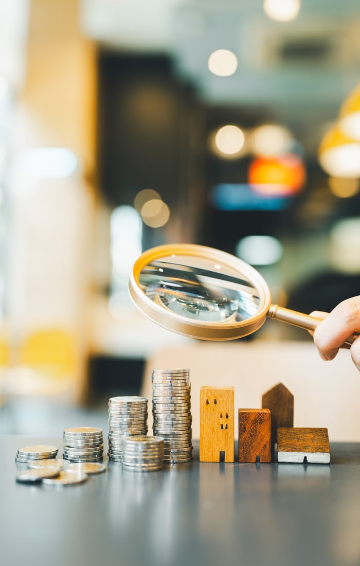 Hand hält Lupe über mehrere Stapel hoch Münzen und Mini Figuren von Holzhäusern